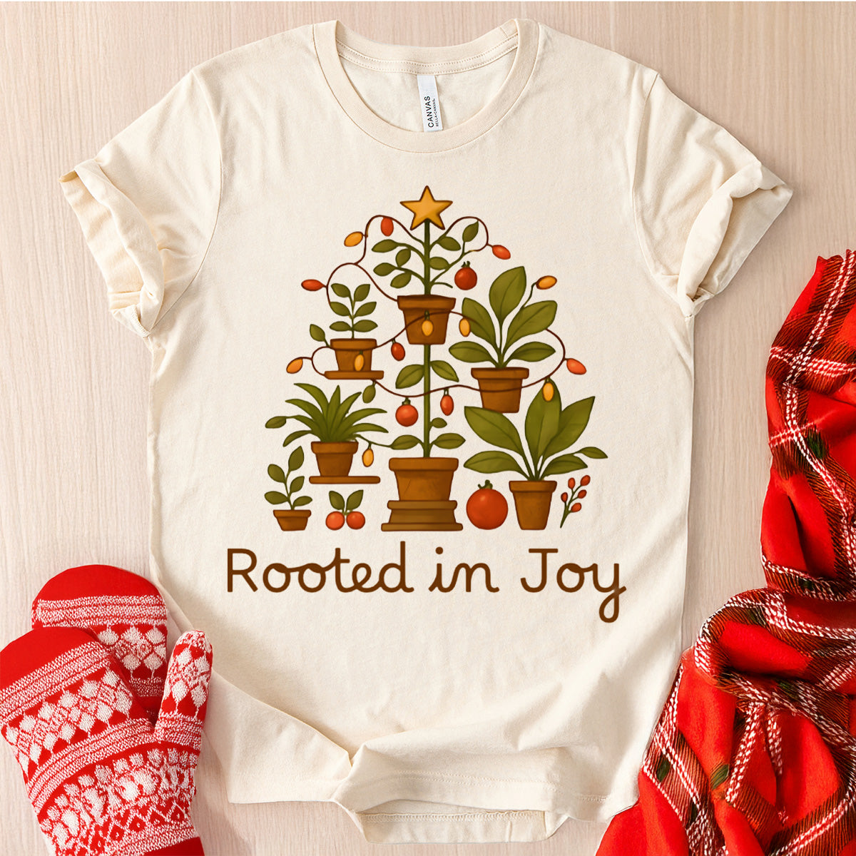 White t-shirt with a Christmas-themed design and 'Rooted in Joy' text, surrounded by festive items on a wooden surface.